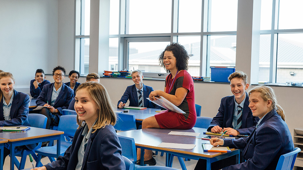 Teacher sitting in a class with students. They are all smiling.